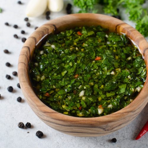 A wooden bowl wirh homemade fresh chimichurri sauce standing on a wooden board with ingredients